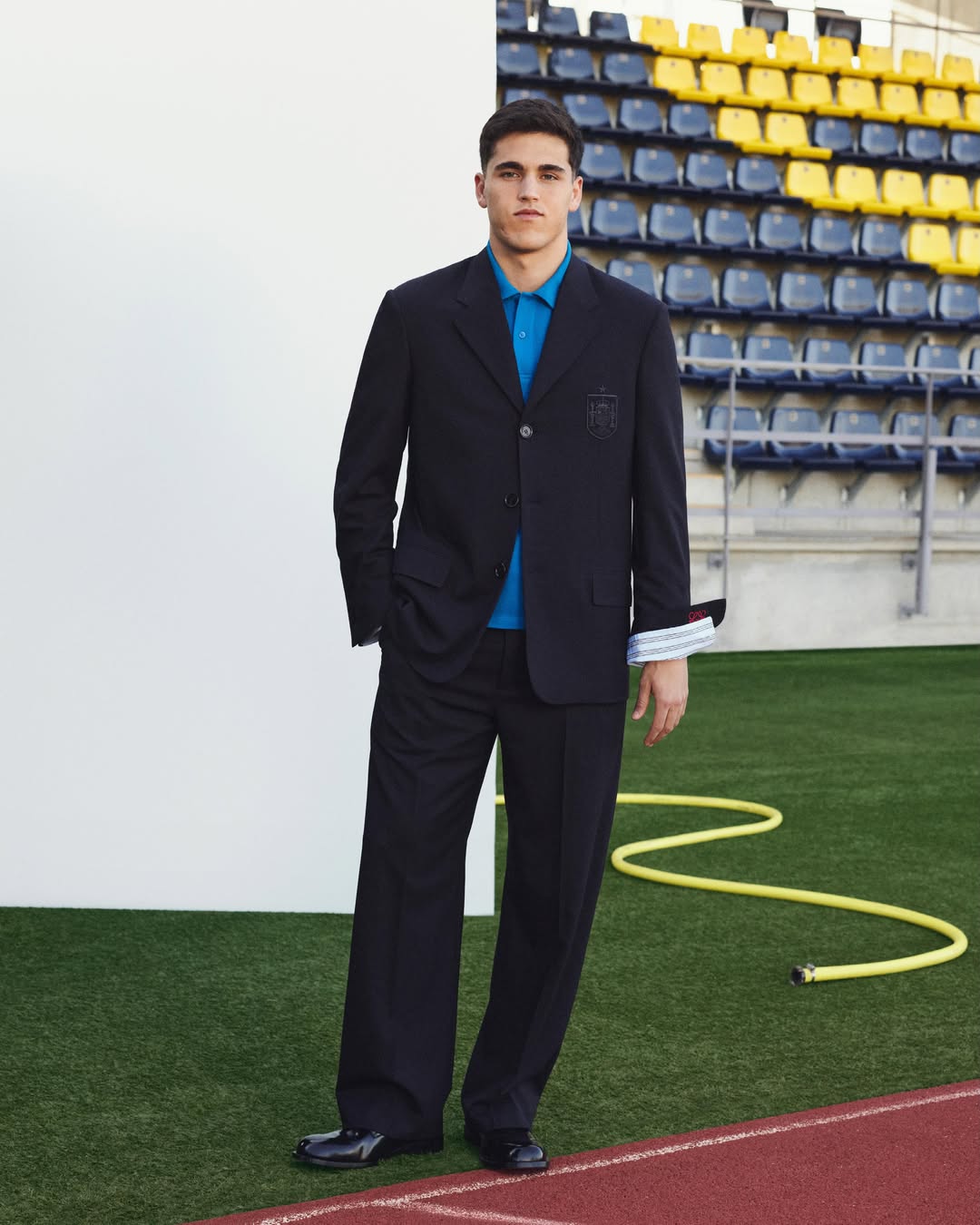 Full-length Loewe x Spain travel wardrobe: navy blazer with the Spanish federation crest, blue polo and wide-leg trousers on a stadium pitch.