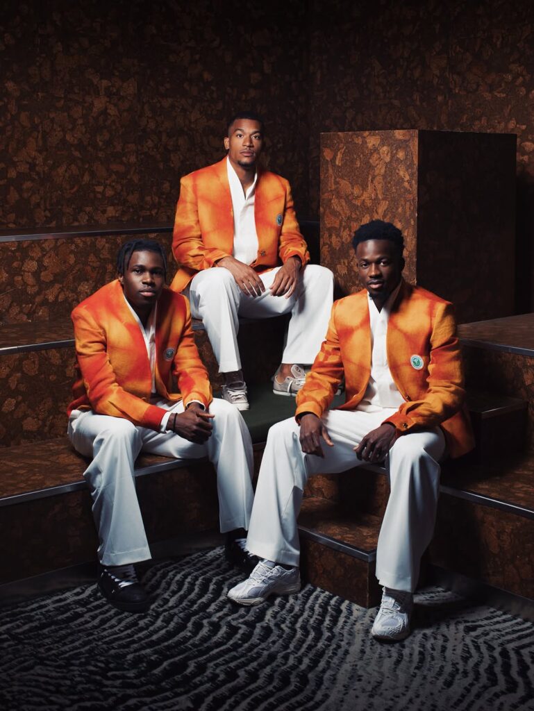If This. Then That: World Cup Edition — Dressing the Team — Loewe → Ibrahim Fernandez 3 Models in Ibrahim Fernandez's saffron-orange ceremonial jackets and white trousers, the Côte d'Ivoire Elephants' off-pitch departure look, posed against a rust-toned backdrop.