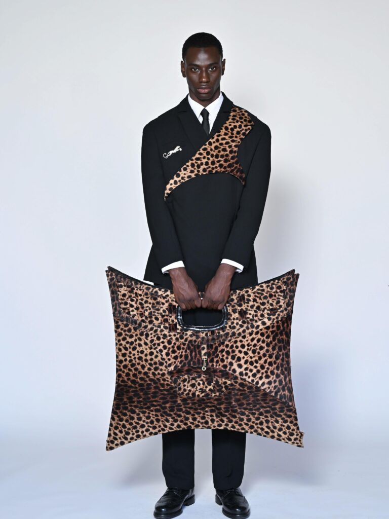 A model wearing the JMAKxPARIS black suit with a leopard-print sash and gold leopard pin, holding the matching leopard-print star bag