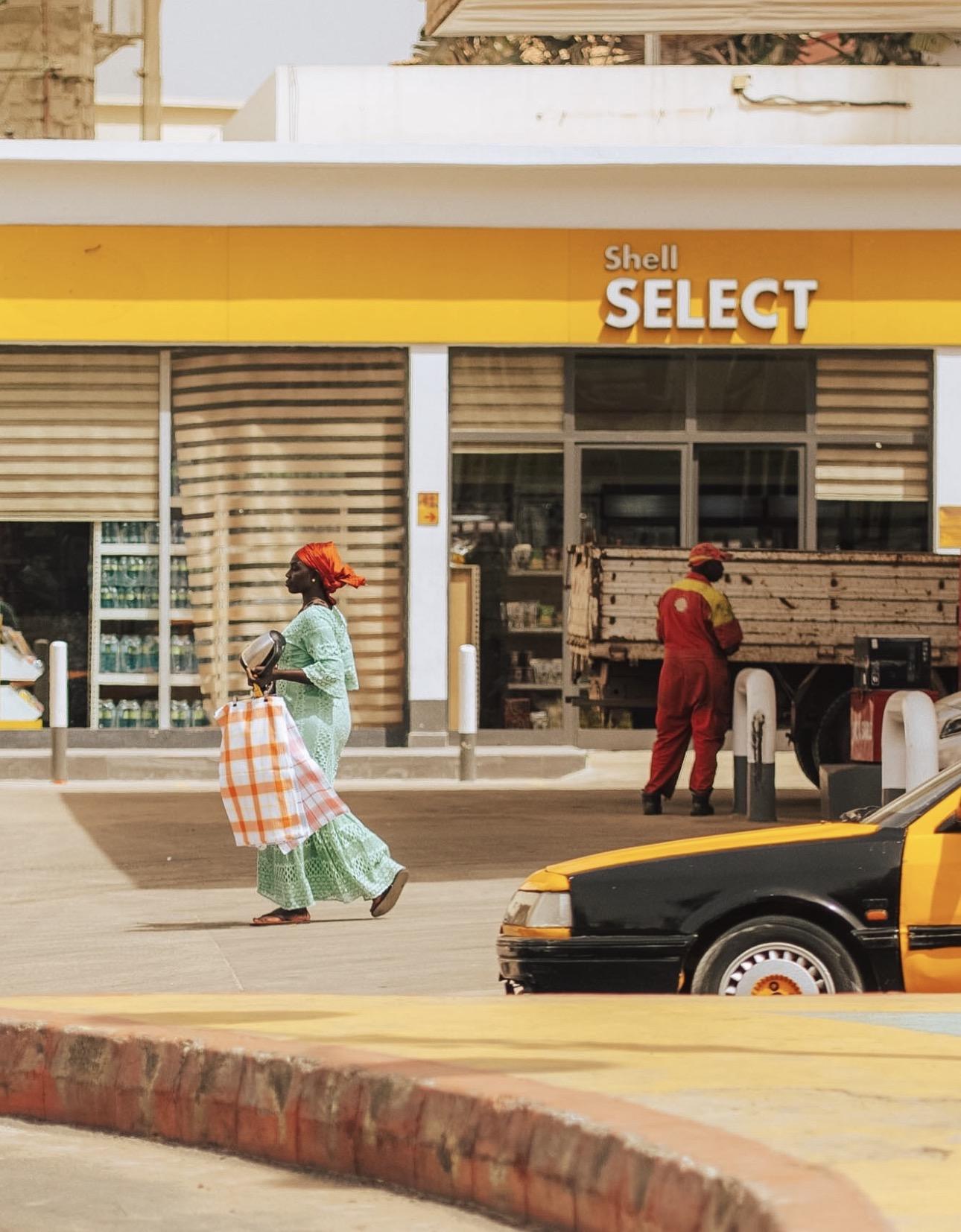 A woman in a green outfit carrying a wrapped bundle walks past a Shell petrol station and a yellow taxi on a Dakar street