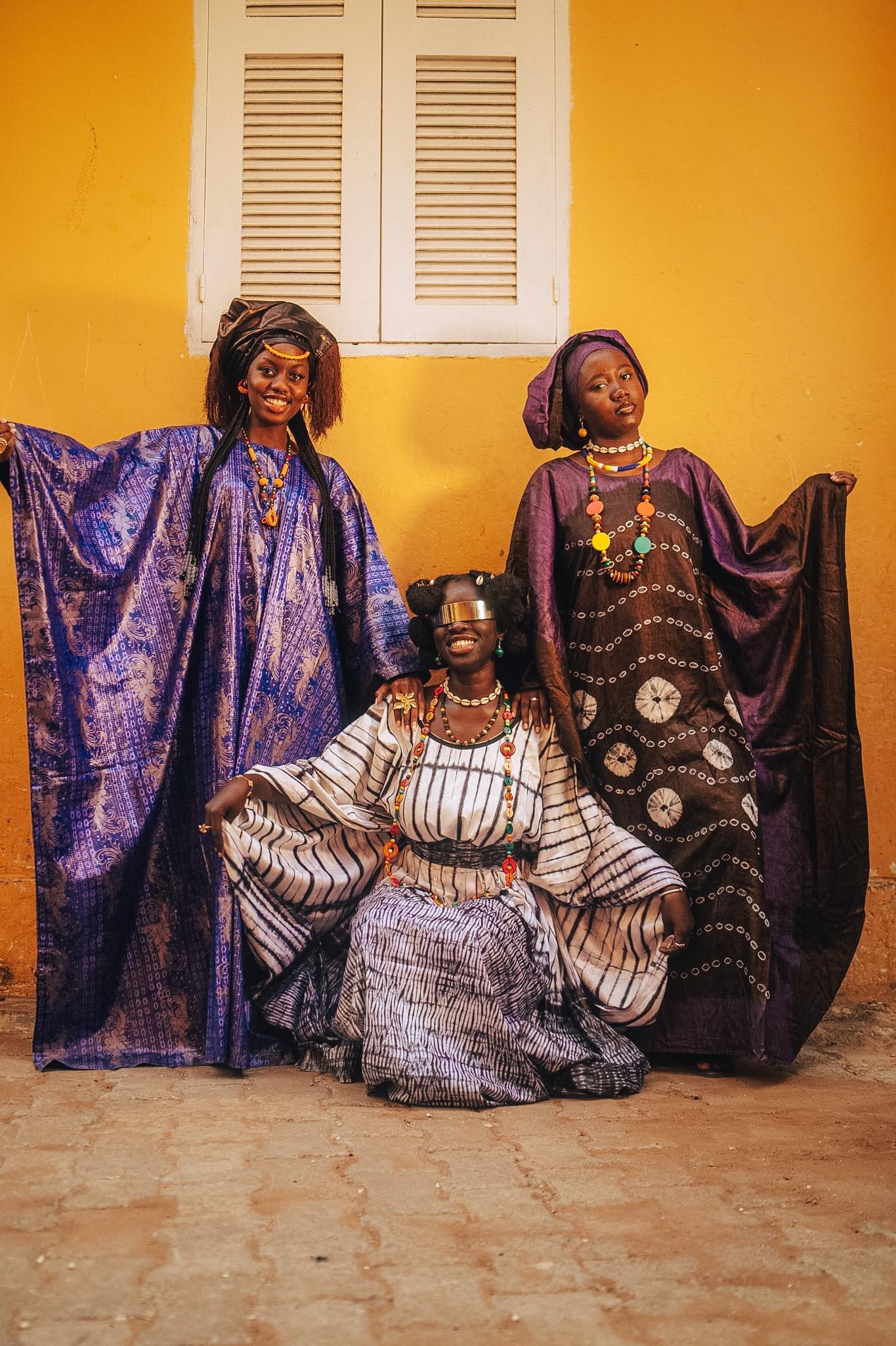 Three women in purple boubous and patterned indigo robes seated and standing against a yellow wall in Dakar