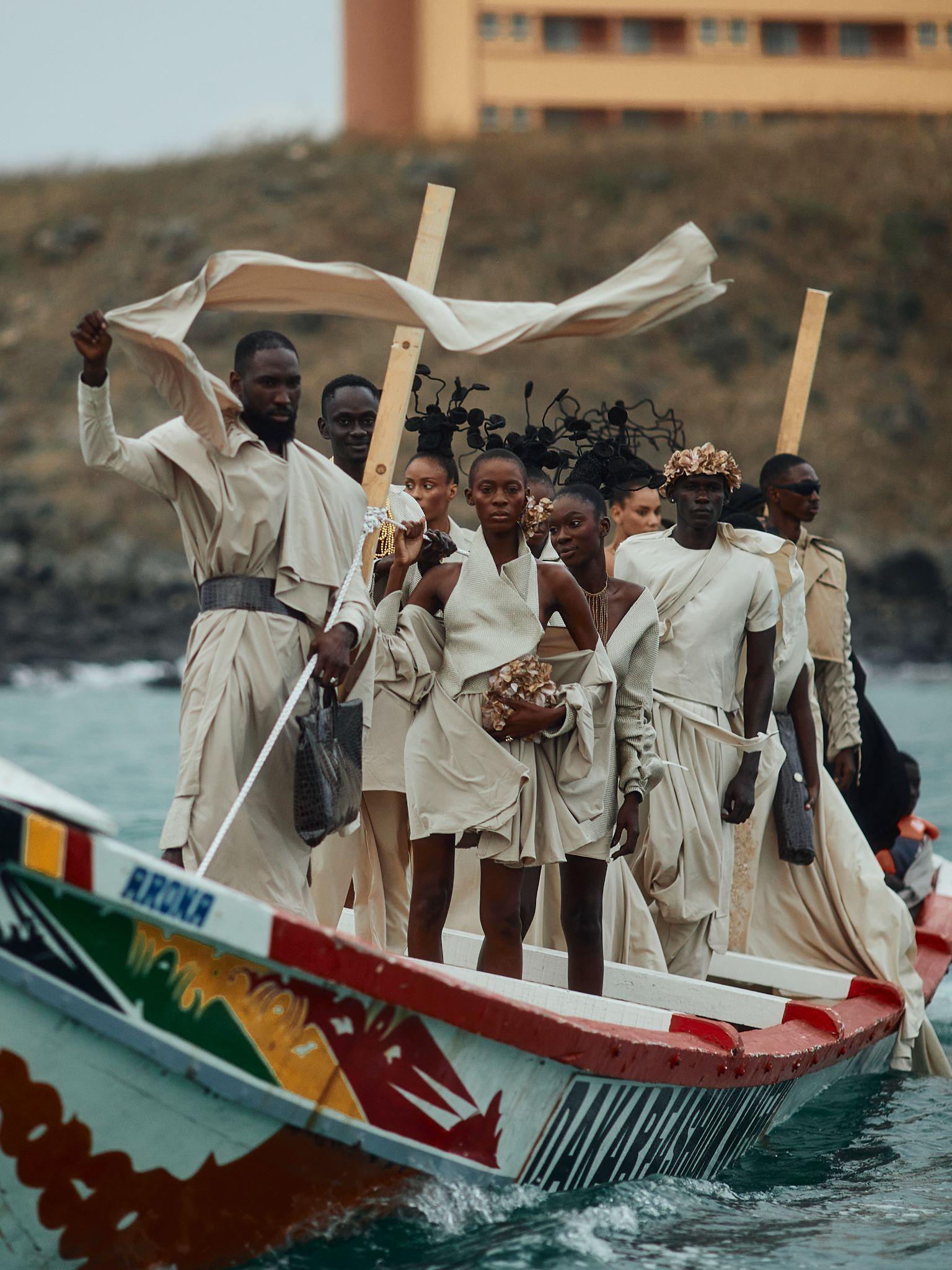 Models in flowing white garments standing in a painted pirogue at sea for Dakar Fashion Week 2025