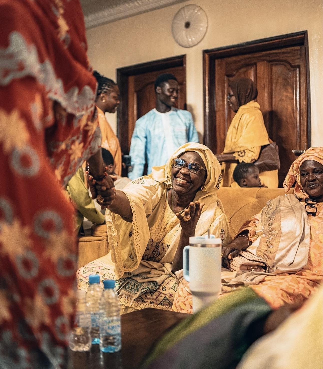Women in embroidered boubous laughing together at a gathering in Dakar, teapots and bottles on a low table