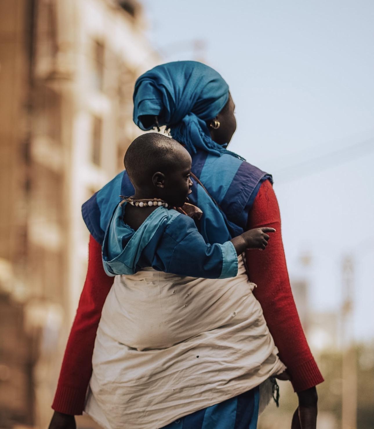 A woman in a blue headscarf carries a child wrapped on her back as she walks a Dakar street on the city's creative economy