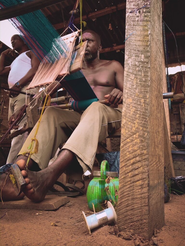Two Yoruba weavers work at traditional narrow-strip looms weaving aṣọ-òkè in an open-air workshop, with warp threads stretched out and spindles of thread on the ground.