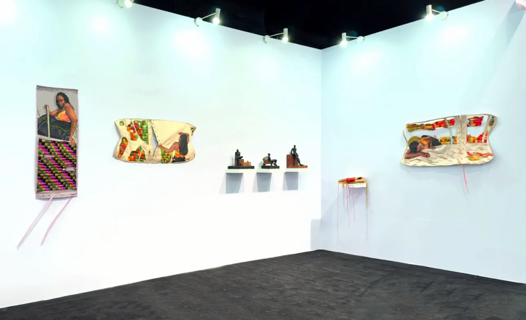 Afriart Gallery booth at ART X Lagos, textile and beadwork panels on white walls with three small figures on wall shelves