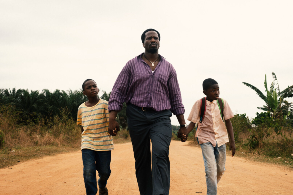 A still from My Father’s Shadow, a man in a striped shirt walking a red dirt road holding hands with two boys
