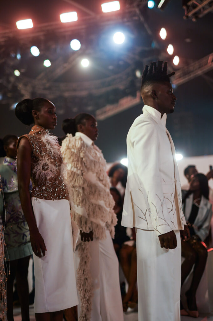 The Money Is the Wrong Shape 2 Models in white embellished eveningwear and feathered detailing lined up under bright stage lights at an African runway show.
