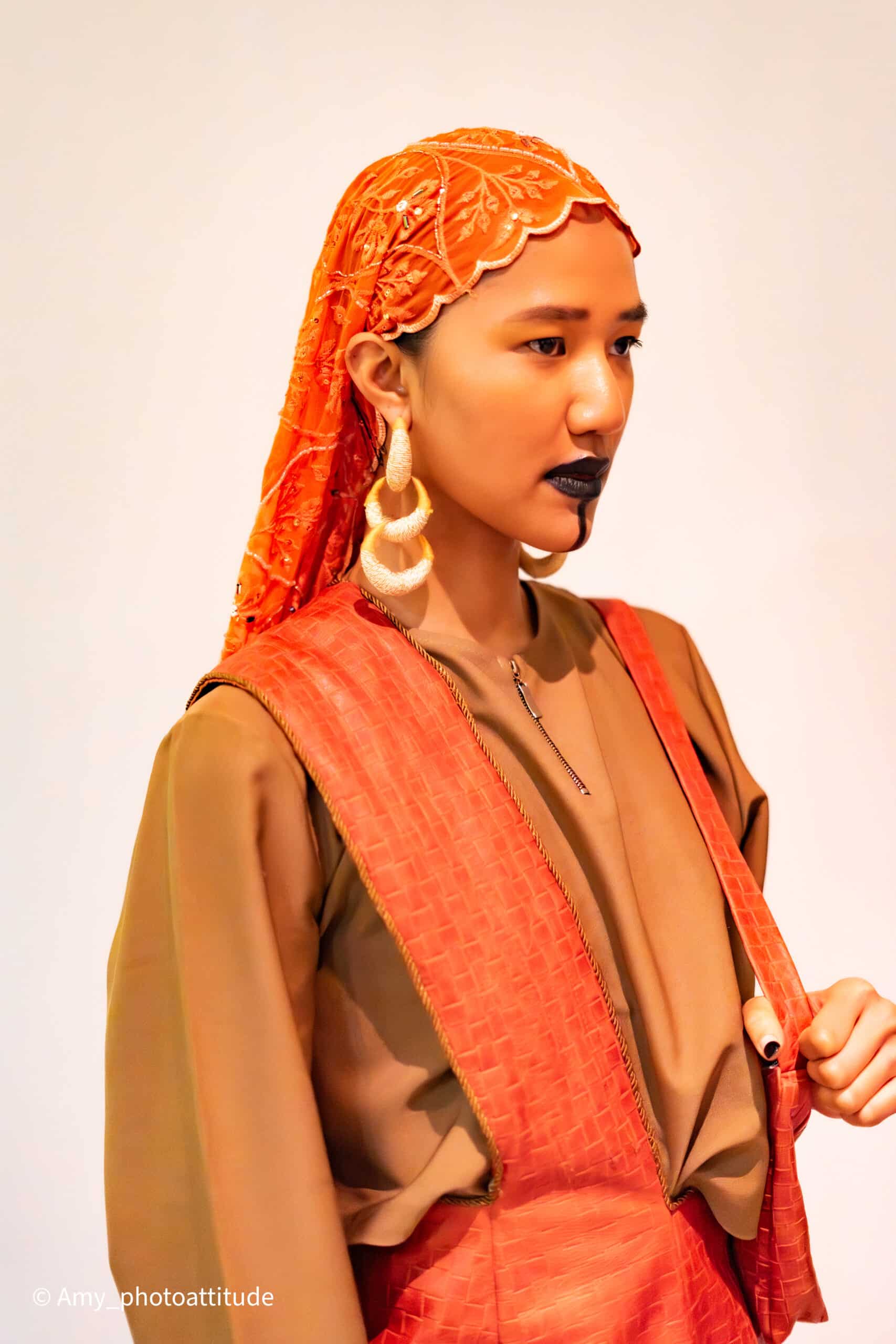 NAANIYA SS26 Tombouctou collection portrait: model in a rust-brown robe with a red patterned sash and hooped earrings at Paris Fashion Week