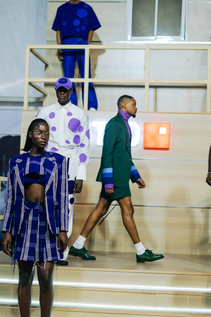 The Money Is the Wrong Shape 3 Models in cobalt-blue plaid and purple polka-dot contemporary looks walking a staircase runway at an African fashion show.