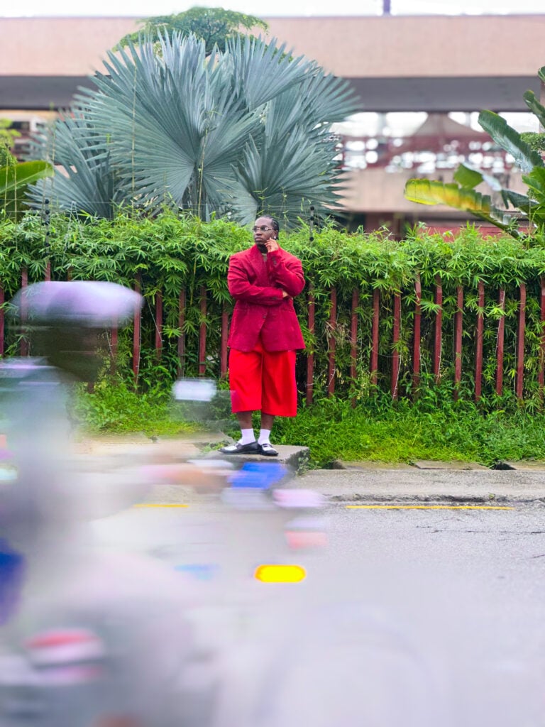 Ugonna-Ora Owoh in a red jacket and red wide-leg trousers on a Lagos kerb, a passer-by blurred in the foreground