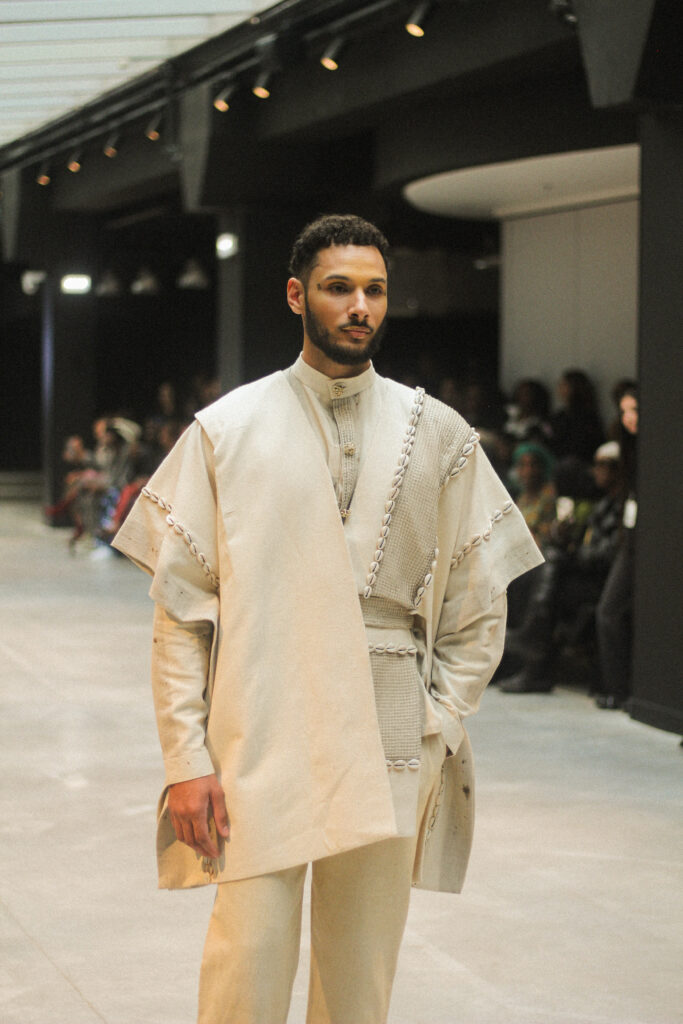 Male model in a cream caped tunic ensemble with braided trim, NAANIYA SS26 Tombouctou collection at Paris Fashion Week