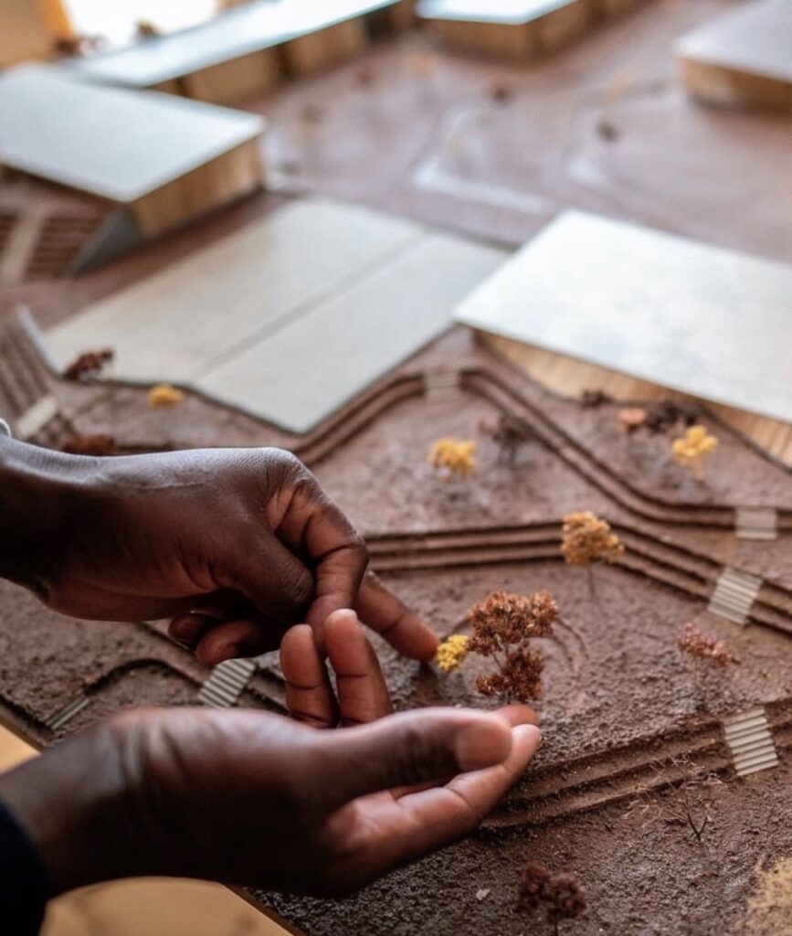 Hands arranging trees on the laterite architectural scale model of the Goethe-Institut Dakar