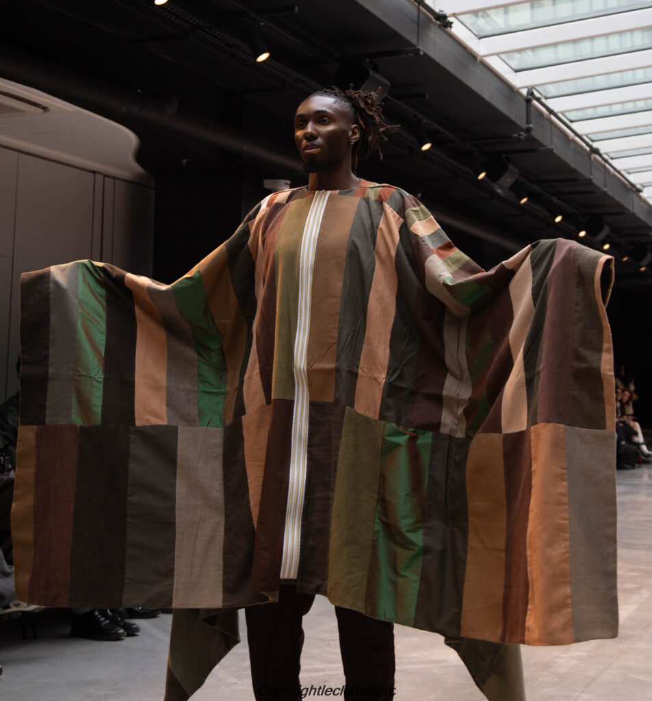 Model presenting a patchwork panel coat in earth tones with arms outstretched, NAANIYA SS26 Tombouctou collection