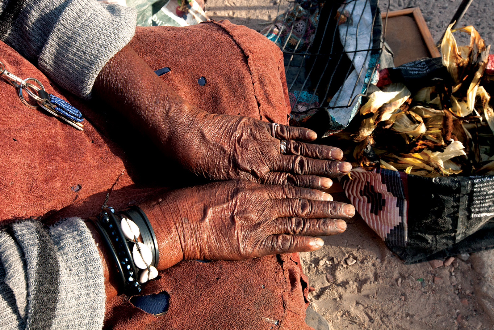Close-up of an older woman's weathered hands resting in her lap, wearing black bangles with cowrie-shell accents, in Bairro 6 de Maio, Amadora.