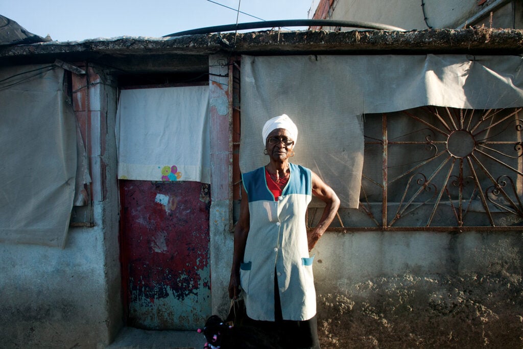 An older woman in a checked apron and white headwrap stands outside her makeshift home in Bairro 6 de Maio, Amadora.