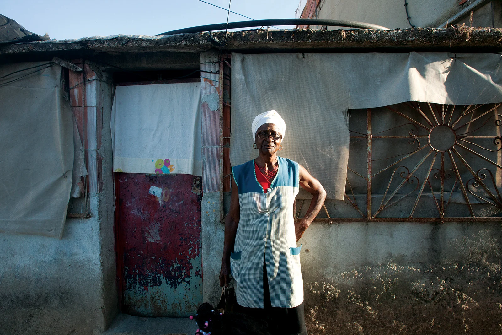 An older woman in a checked apron and white headwrap stands outside her makeshift home in Bairro 6 de Maio, Amadora.