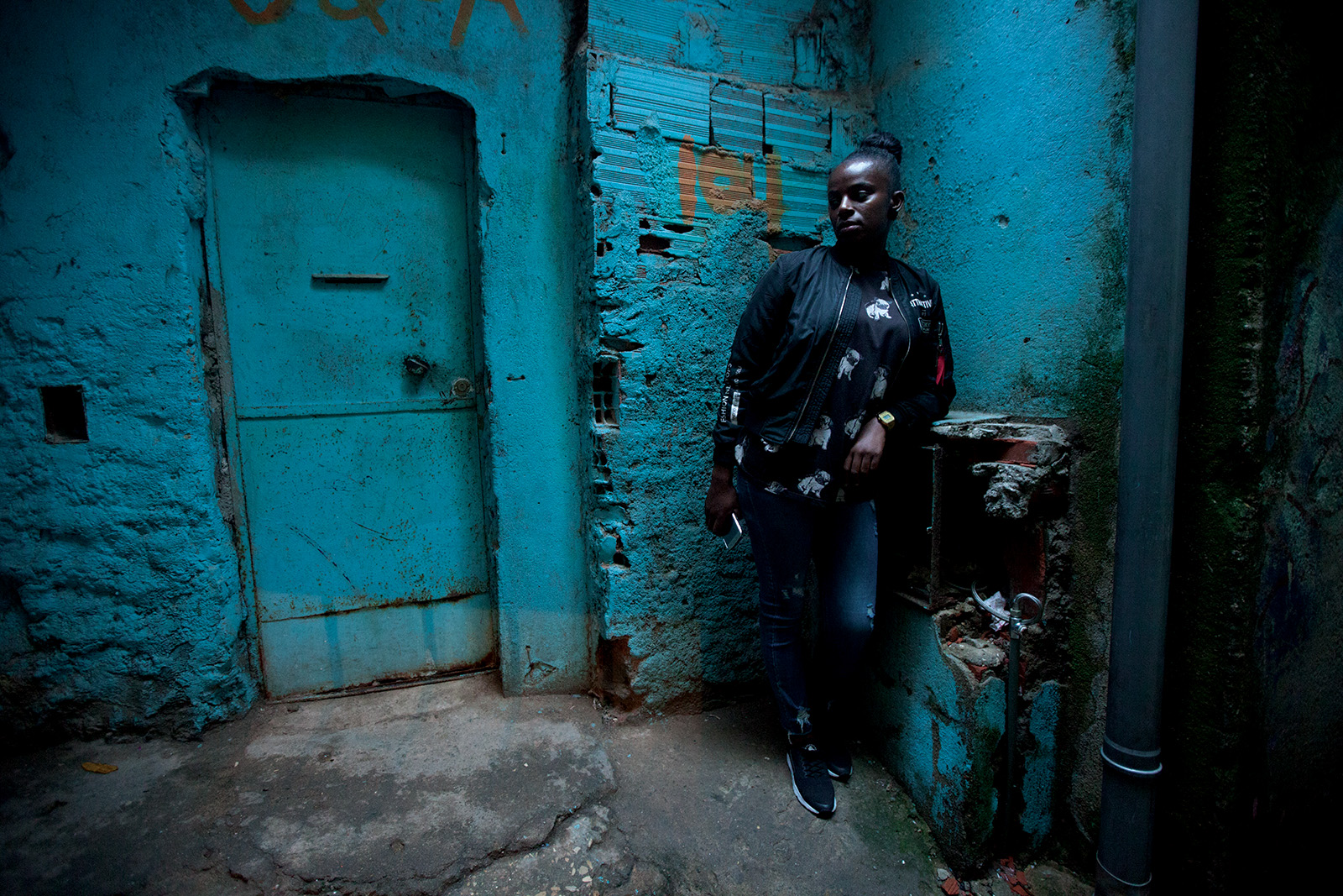 A young woman leans against a teal weathered wall in a doorway of Bairro 6 de Maio, the Cape Verdean neighbourhood near Lisbon.