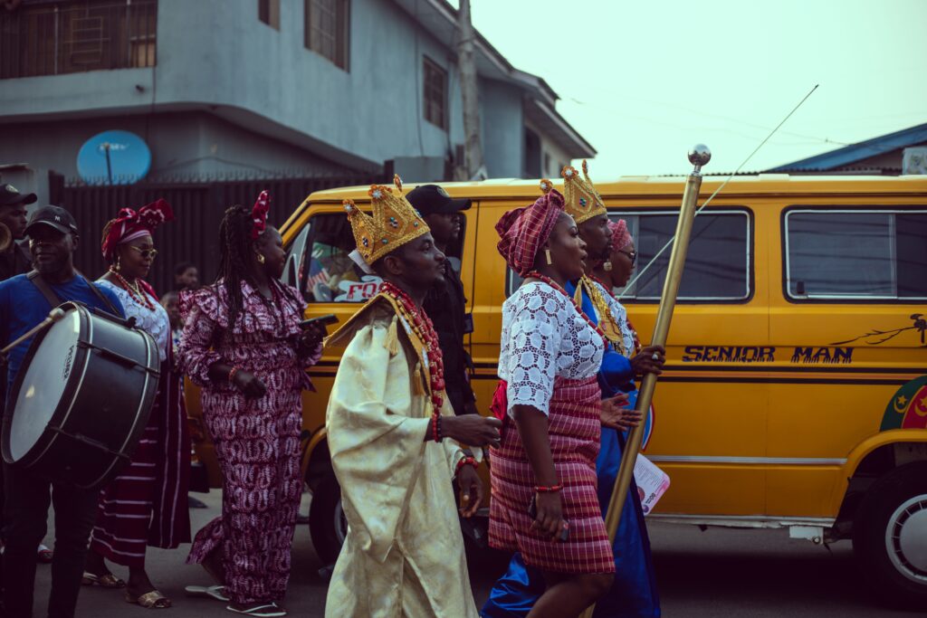 A Lagos street procession in gold crowns and coral beads passing a yellow danfo bus, a bass drummer at the left