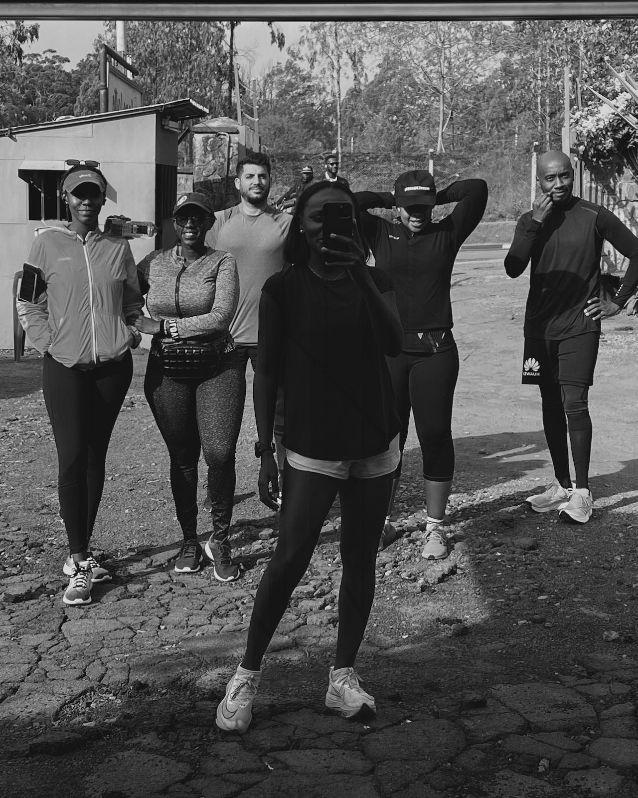 Black-and-white photograph of six runners standing on a road, one in a dark hood in the foreground.