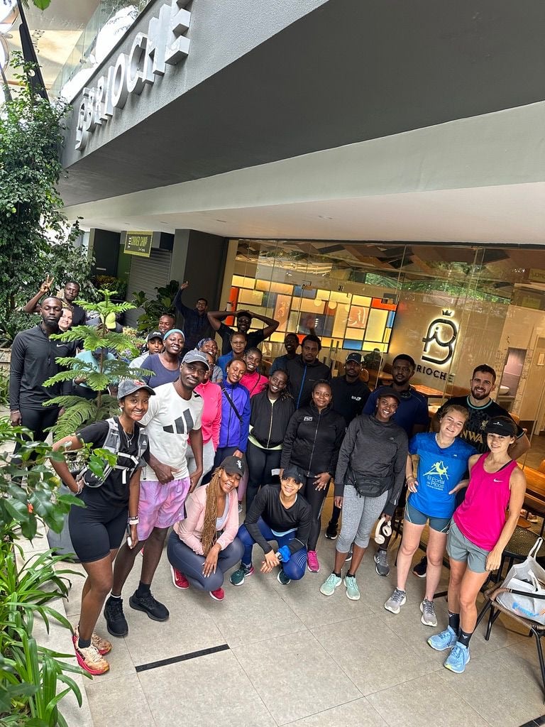 About twenty runners gathered for a photograph outside a cafe under a concrete overhang, planting in the foreground.