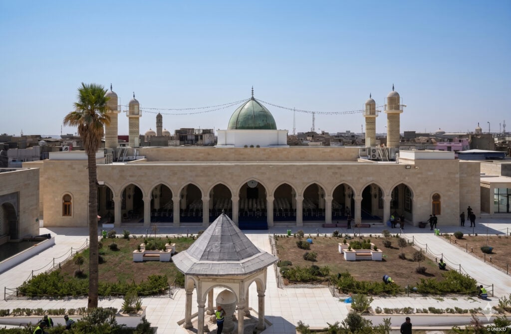 The rebuilt Al-Nouri complex in Mosul from above: an arcaded stone courtyard, green-domed prayer hall, two minarets and replanted gardens against the city skyline.