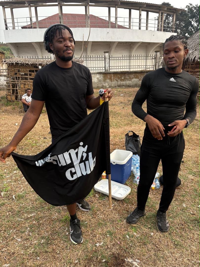 A man holds a black club banner open beside a teammate in black, a cool box and a curved concrete grandstand behind them.