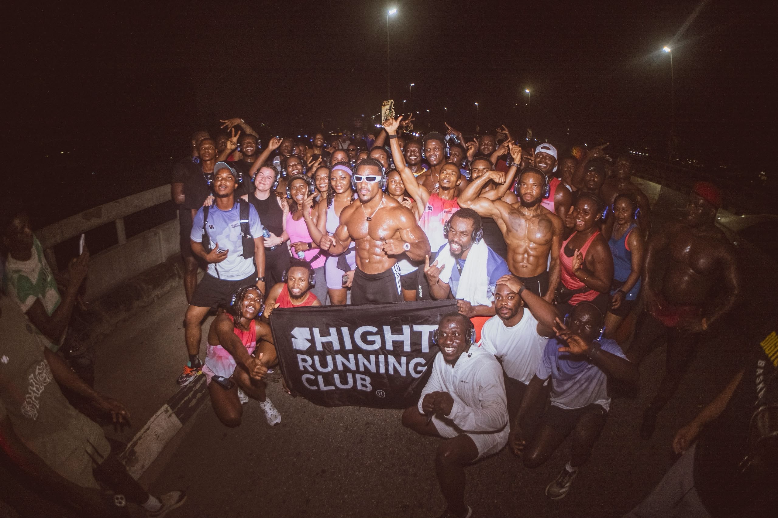 A large night-time crowd behind a club banner, a man at the centre with both arms raised under warm light.