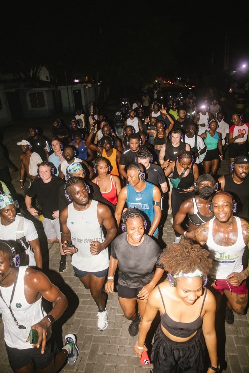 A dense crowd of night runners moves toward the camera wearing headphones, faces lit by camera flash.