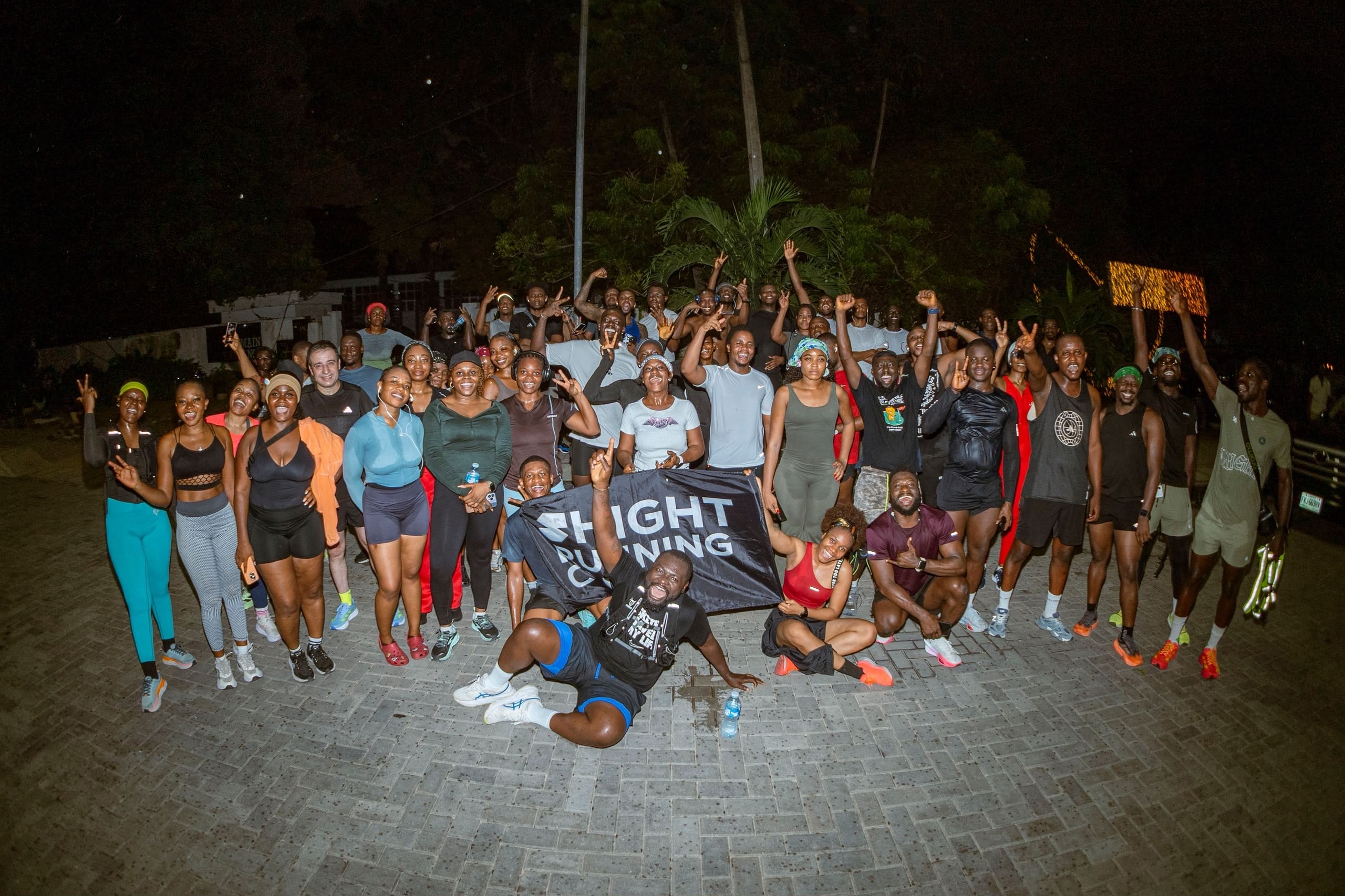 More than fifty runners pose at night on a Lagos road, arms raised behind a club banner, lit by a single streetlight.