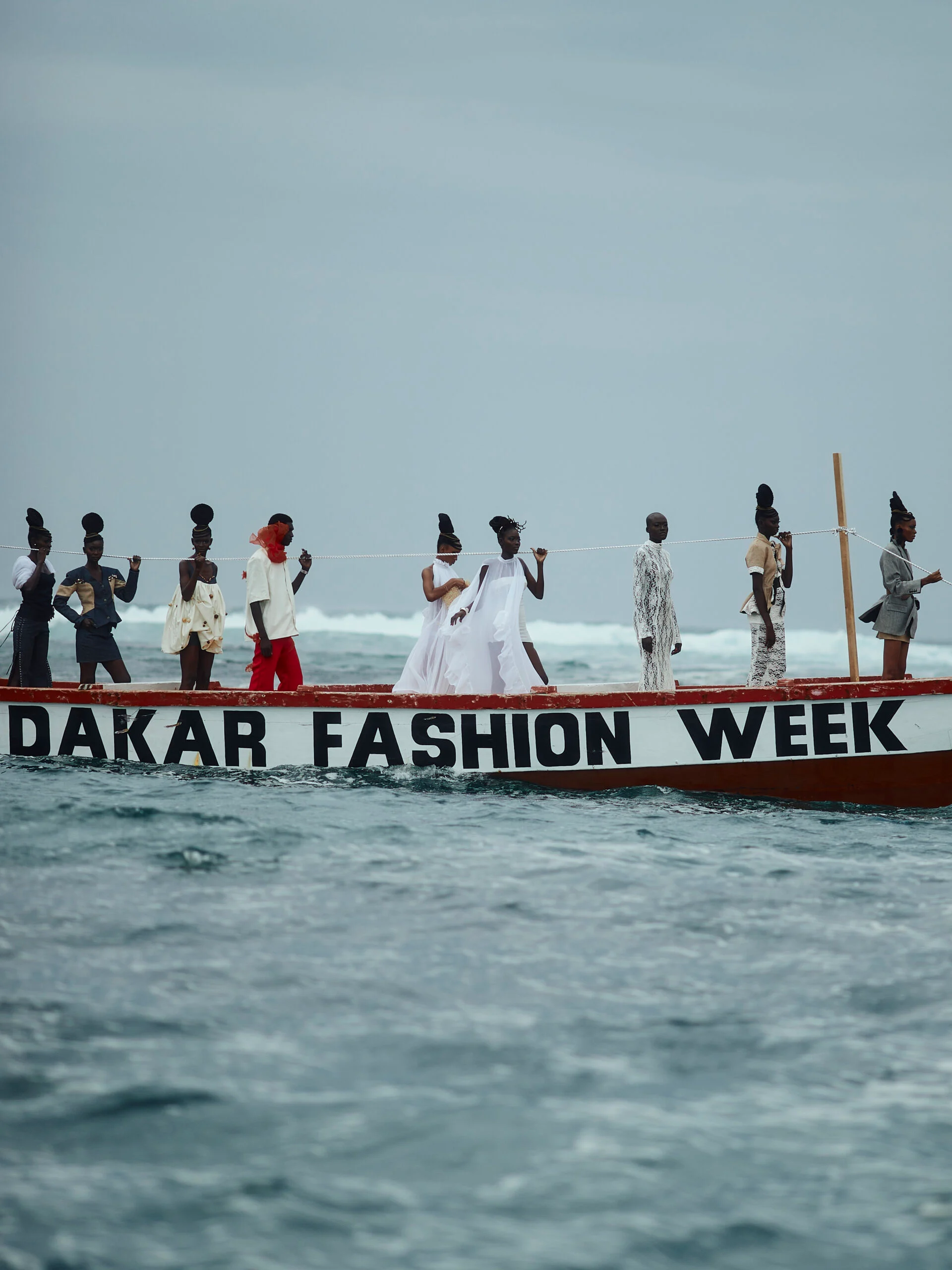 Ten models stand along a fishing pirogue painted with the words Dakar Fashion Week, on grey open water under an overcast sky.