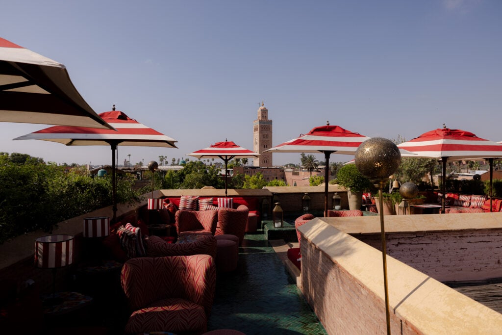 A rooftop terrace at El Fenn, Marrakech, with red-and-white striped parasols and low seating, the Koutoubia minaret on the skyline beyond.