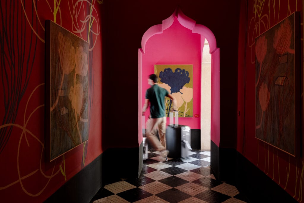 A pink-walled alcove seen through a keyhole arch at El Fenn, Marrakech, with a figure beside framed art and a black-and-white tiled floor.