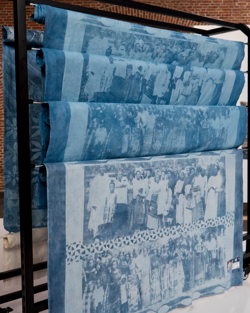 Adirẹ and the Wrapper-Wearers 1 Indigo cloth panels printed with archival photographs of Nigerian women, draped over a black metal clothing rack in a brick-walled hall.