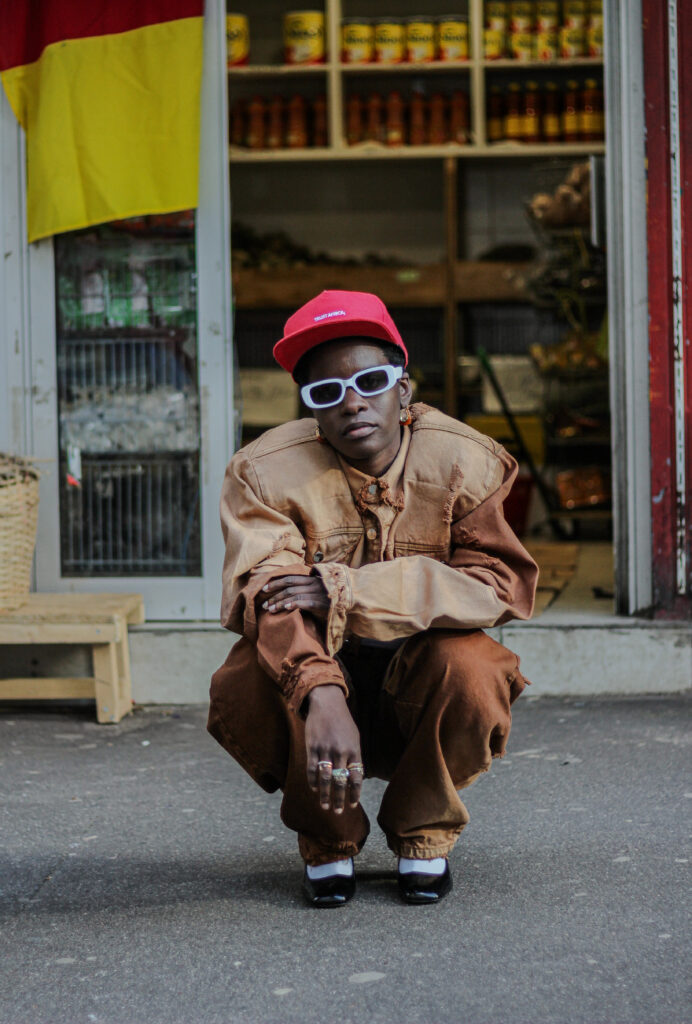 Bledard; Seris A model in a tan workwear set, red cap and sunglasses crouching outside a shopfront