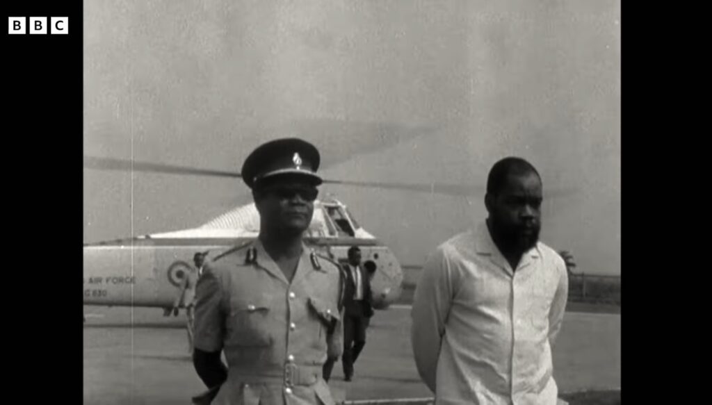 Chukwuemeka Odumegwu Ojukwu, bearded and in a light shirt, walking across a tarmac beside a uniformed officer in front of a Nigerian Air Force helicopter.