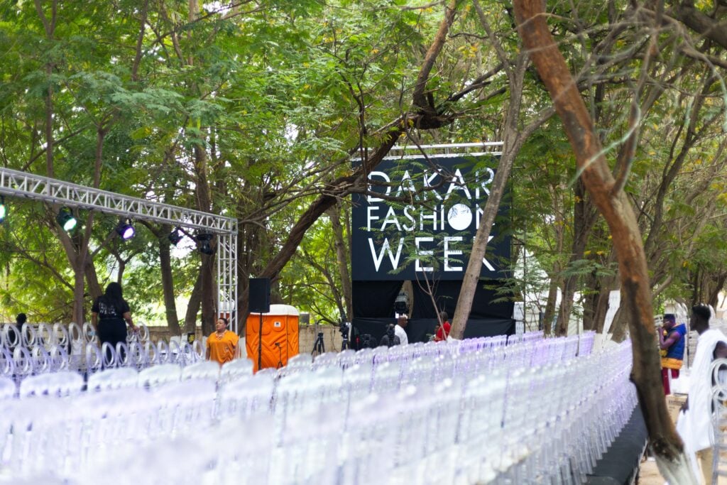 An open-air runway set among trees at Parc de Hann, with a lit stage backdrop reading Dakar Fashion Week and rows of clear chairs in the foreground, Dakar, 2024.