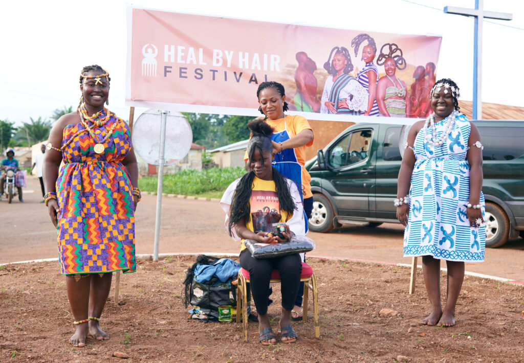 Heal by Hair: How Communities, Culture, and Trust Are Redefining Mental Health Across Africa 1 Three women in wax print dresses stand beneath a Heal by Hair Festival banner in Siou, Togo, while a hairdresser styles the hair of a woman seated on a stool