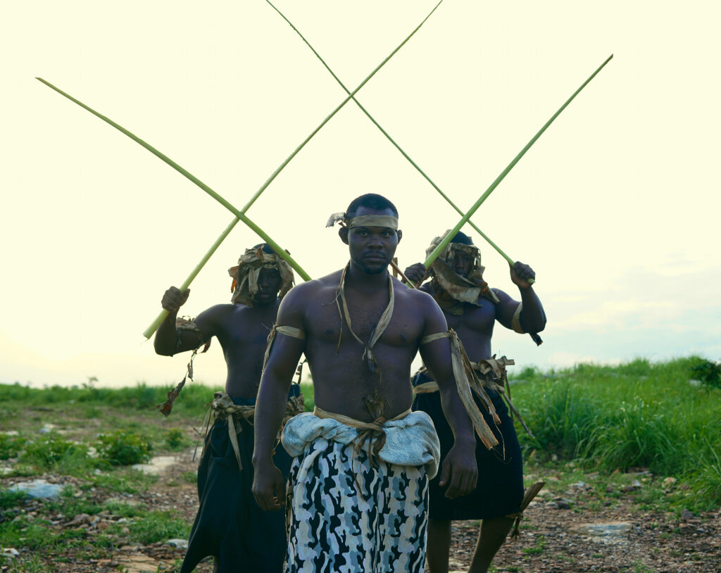 Heal by Hair: How Communities, Culture, and Trust Are Redefining Mental Health Across Africa 3 Bare-chested wrestlers of the Cameroonian Ngondo delegation walk through a grass field beneath raised cane stalks at the Heal by Hair Festival in Siou, Togo