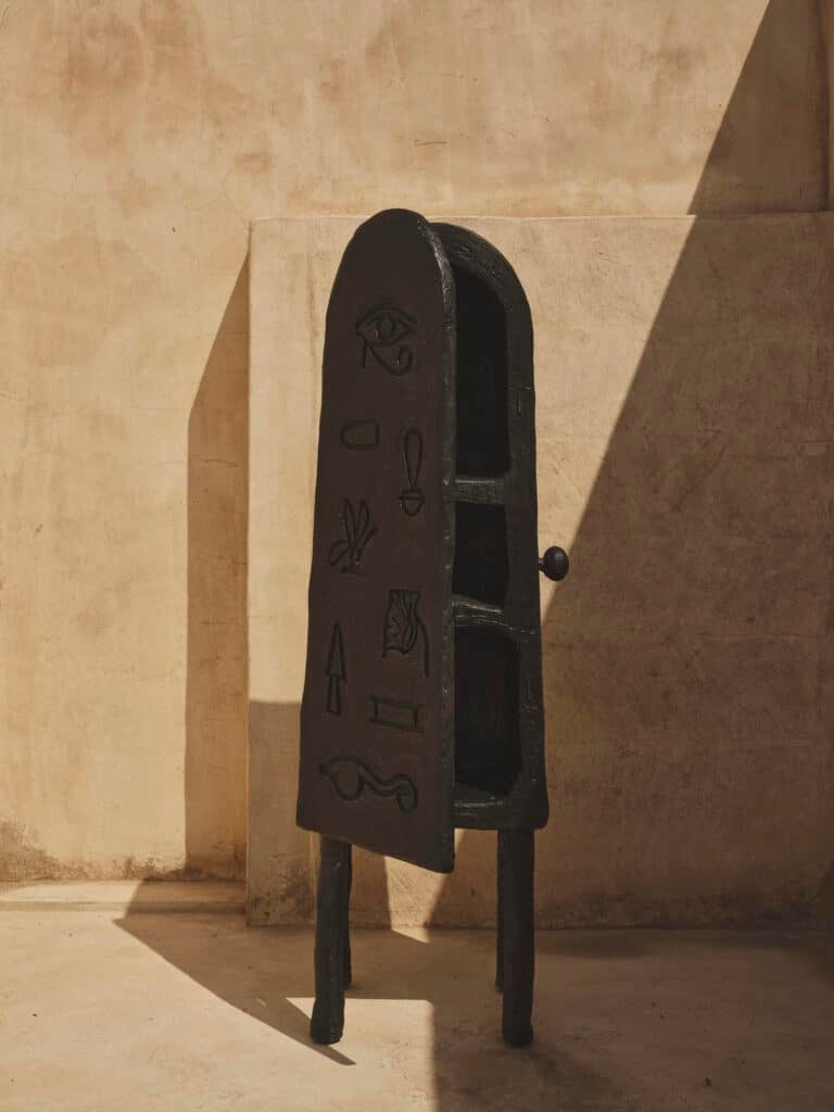 Byblos, a tall arch-topped cabinet in dark carved wood with incised panels, standing in raking sunlight against a plaster wall.