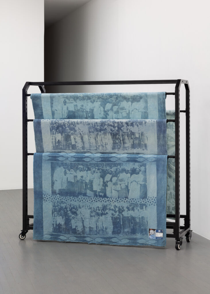Adirẹ and the Wrapper-Wearers 6 Indigo cloths carrying archival group portraits of women, hung over a black rolling garment rack in a white gallery corner.