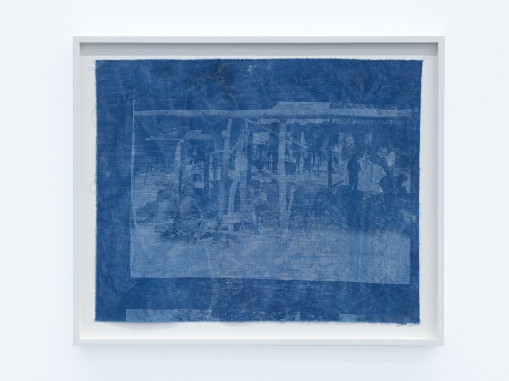 Adirẹ and the Wrapper-Wearers 5 A framed indigo cloth panel showing a faint archival scene of figures working beneath an open timber shelter.