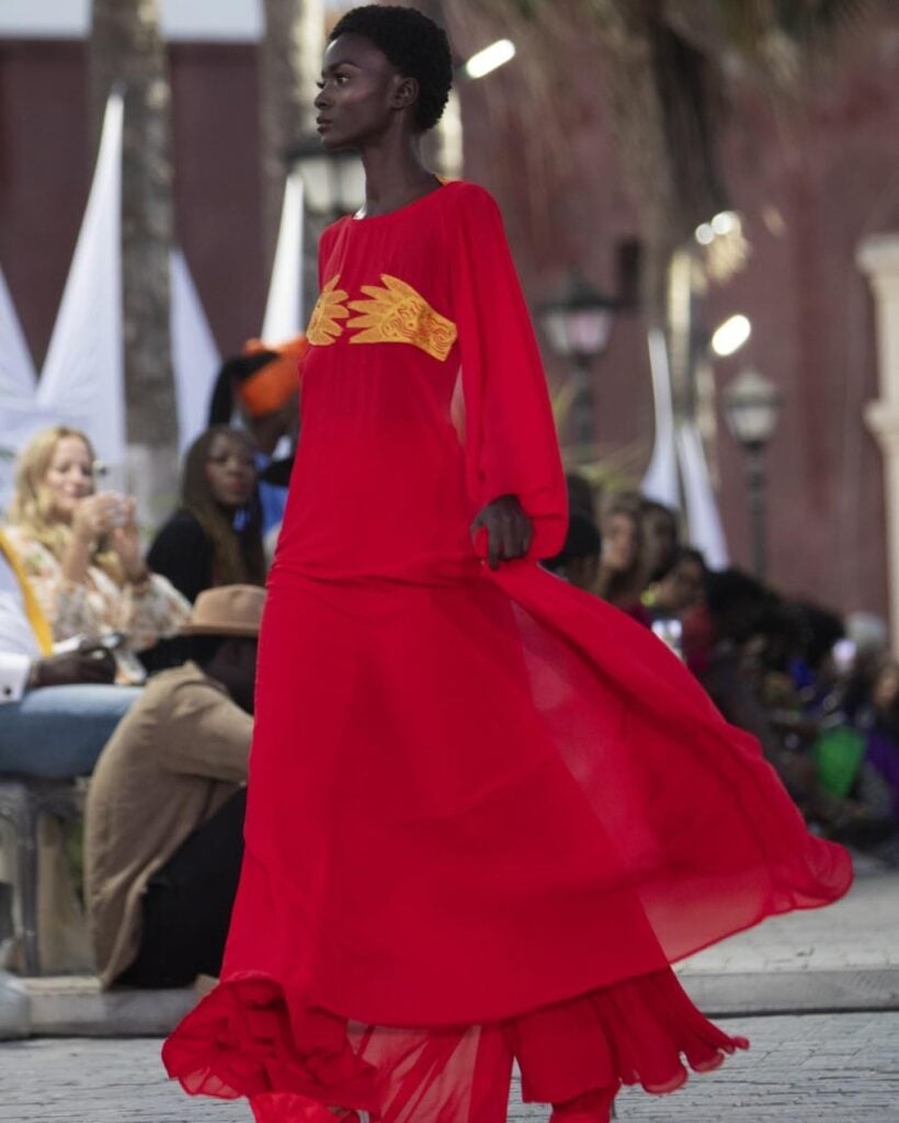 A model in a red gown with gold appliqué walks a cobbled street on Gorée Island past a seated audience, colonial arcades and palms behind her.