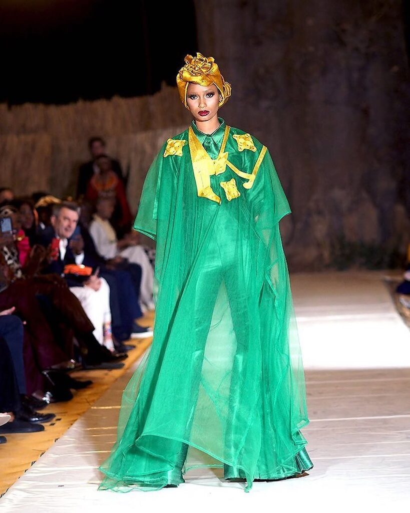A model in a green organza gown with gold appliqué and a matching headwrap walks a white runway among the baobabs near Nguekhokh, Senegal, 2021.