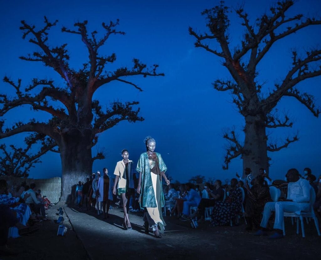 Two models walk a lit path at dusk between two large baobab trees, with a seated audience in deep blue light on either side.