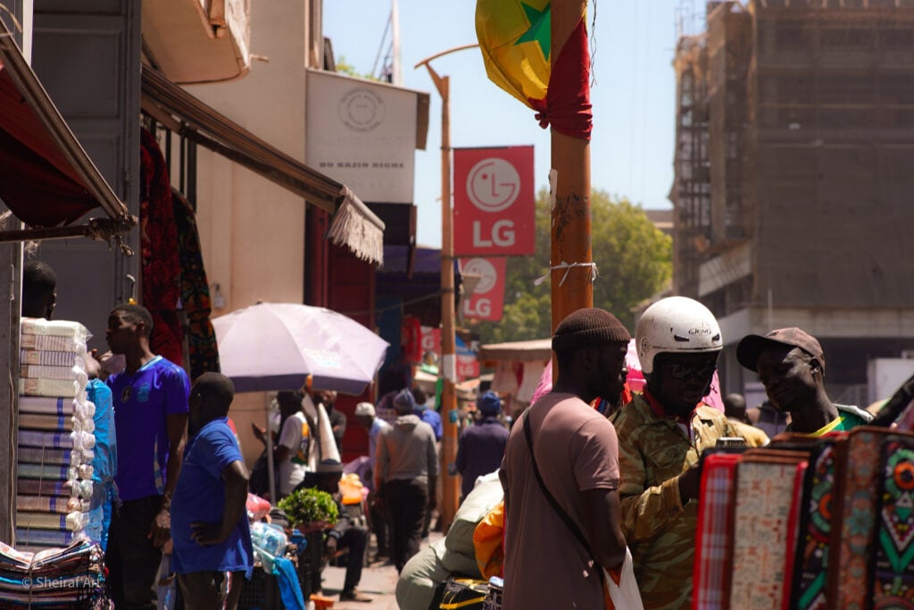 City Codes: Dakar Is Not a Trend 1 Shoppers and fabric stalls along a busy textile market street in Dakar, a Senegalese flag flying above the crowd.