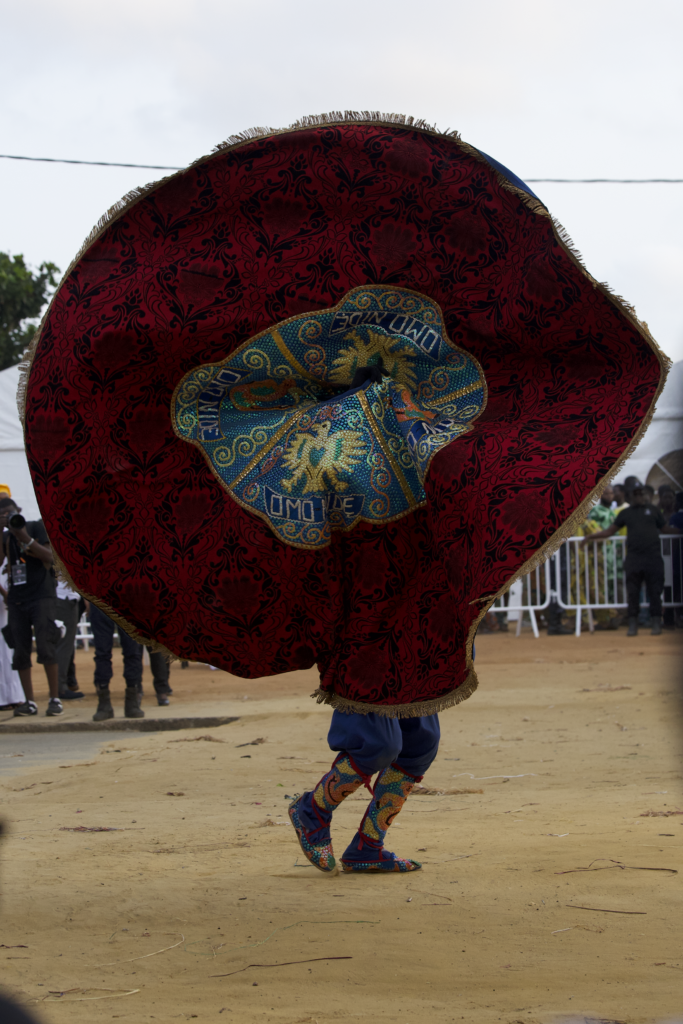 A masquerade opening its beaded cape into a full circle, the central panel reading Omo Nide, filling the street