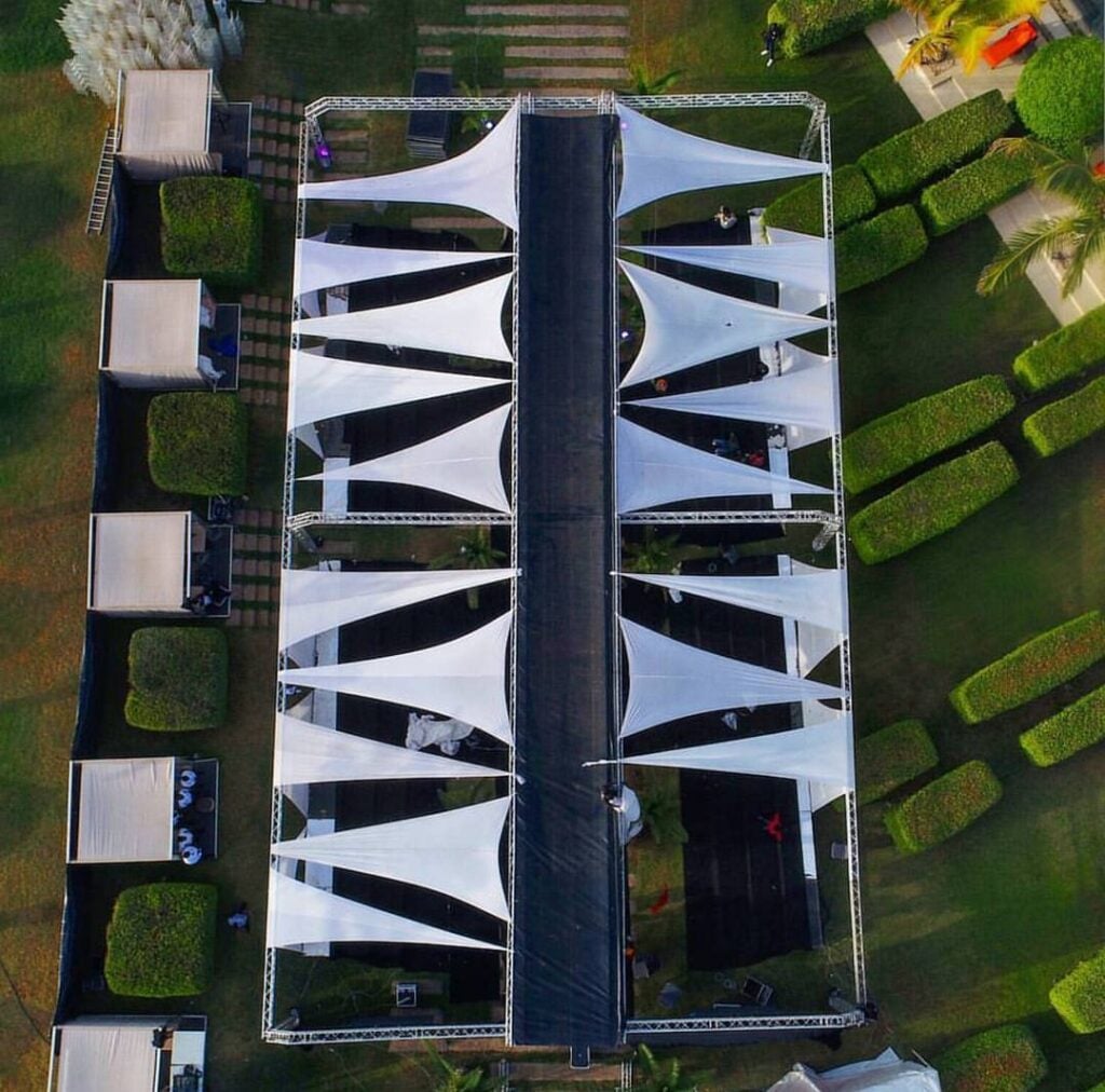 Overhead view of an outdoor runway under white sail canopies rigged on scaffolding, flanked by lawn, hedges and service tents.