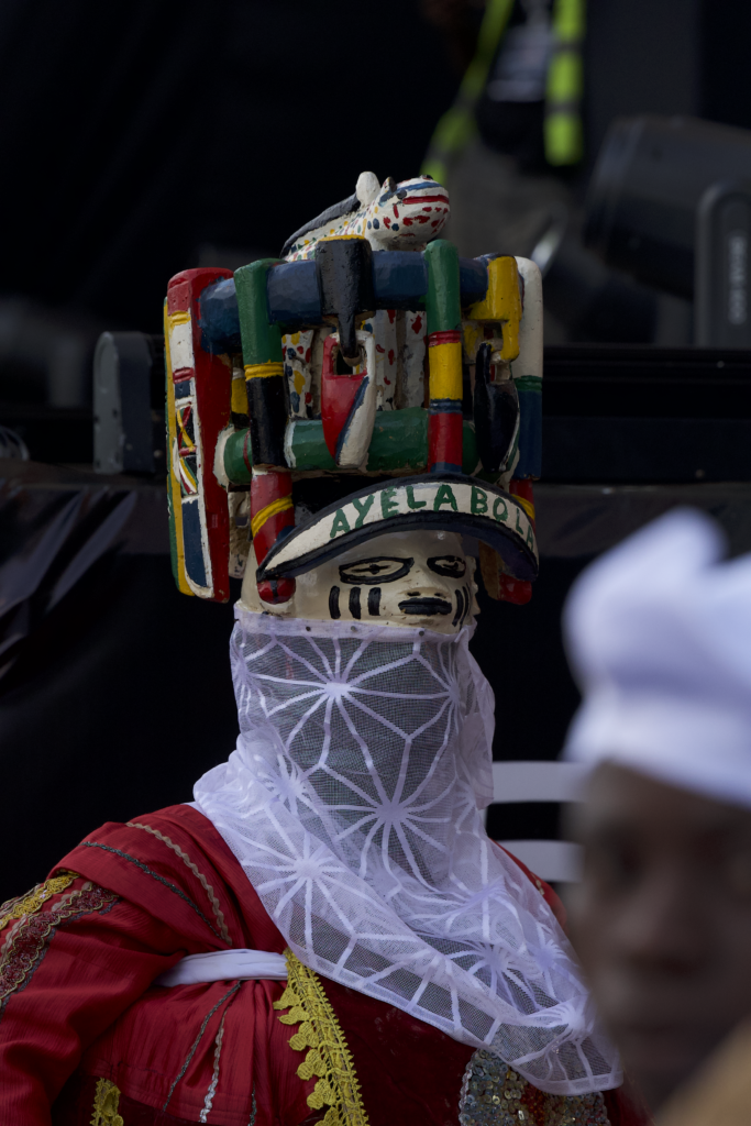 A masquerade named Ayelabola in a carved wooden crest and white lace veil resting between performances