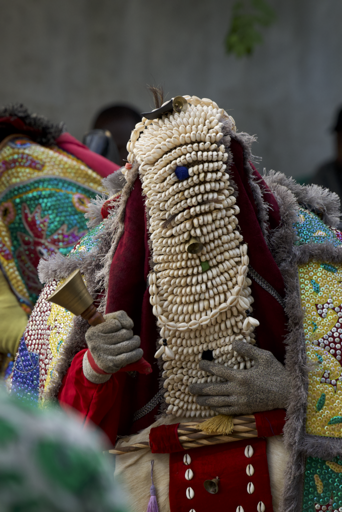 A masquerade with a face covered entirely in cowrie shells holding a brass bell in a gloved hand